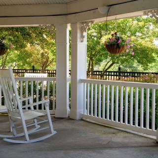 A serene porch with a white rocking chair and hanging plants overlooks a sunny garden.