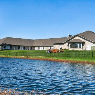 A beautiful house with a large pond in the foreground, surrounded by a well-manicured lawn and a fence under a clear blue sky.