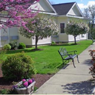 A charming house with a lush garden featuring blooming trees, colorful flowers, and a bench beside a concrete sidewalk.