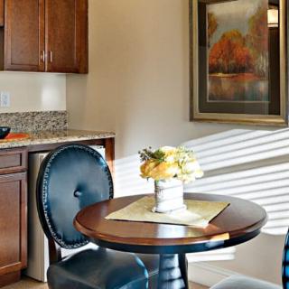 A cozy dining area with a round table, two chairs, and a flower arrangement, next to a kitchen.