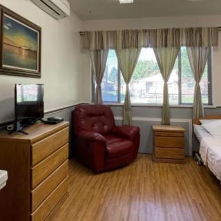 A cozy bedroom featuring a bed, red armchair, dresser with a TV on top, and large windows with curtains.