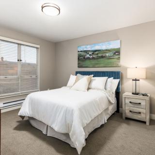 A cozy bedroom with a neatly made bed, a nightstand with a lamp, and a large window providing natural light.