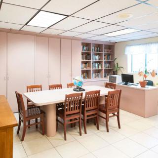 A well-lit office meeting room with a large conference table surrounded by chairs, a computer on the desk, and a bookshelf.