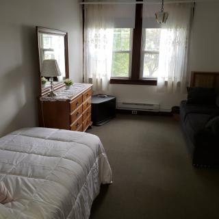 A simple bedroom with a bed, dresser, lamp, and sofa near a window.