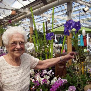 An elderly woman happily stands in a greenhouse surrounded by colorful flowers and plants.