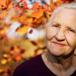 An elderly woman standing outdoors with autumn leaves in the background.