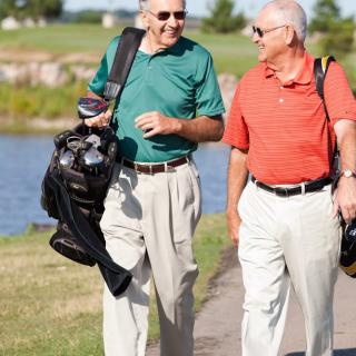 Two senior men walking and chatting on a golf course path, carrying their golf bags.