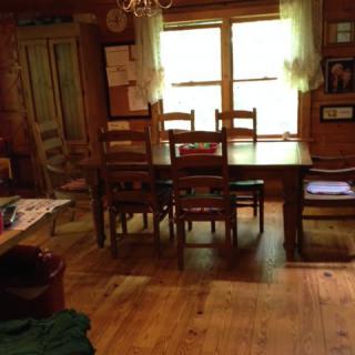 A rustic dining room with a wooden table and chairs, illuminated by natural light from the window.
