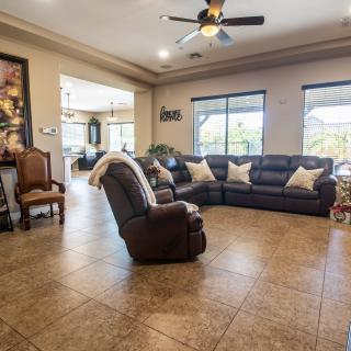 A spacious living room with a sectional sofa and armchair on a tiled floor, featuring large windows and a ceiling fan.