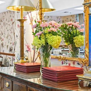 A sophisticated interior decor featuring floral arrangements in vases, gold lamps, and stacked plates on a polished wooden surface with a large ornate mirror.