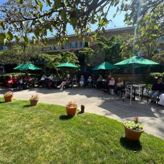 People enjoying a sunny day at an outdoor cafe with green umbrellas and a grassy area.