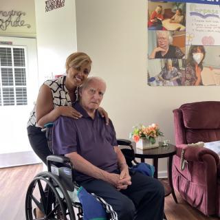 A caregiver smiling while standing behind an elderly person in a wheelchair, set in a cozy home environment.