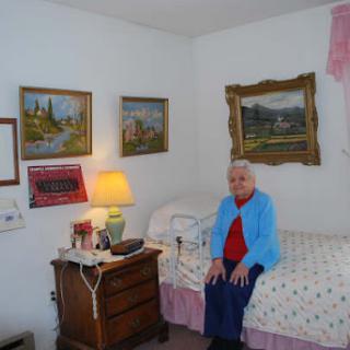 An elderly woman seated in her cozy bedroom, surrounded by paintings and soft lighting.