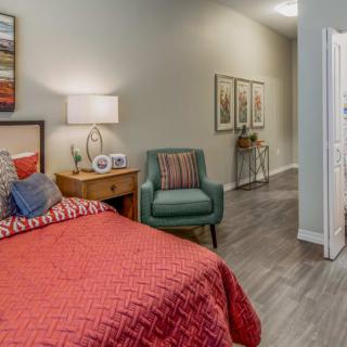A cozy bedroom with a red bedspread, green chair, and a nightstand with a lamp.