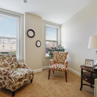 A cozy living room area with two patterned armchairs, a side table with a lamp, and large windows allowing natural light to fill the space.