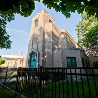 A view of a church building with an art deco design, surrounded by trees and a black iron fence on a clear day.