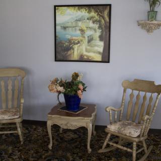 A cozy living room setup featuring two wooden chairs, a table with a vase of flowers, and a landscape painting on the wall.