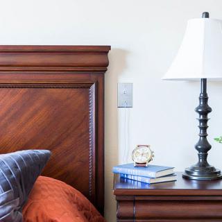 A cozy bedroom setup featuring a wooden headboard, a nightstand with books, an alarm clock, and a lamp.