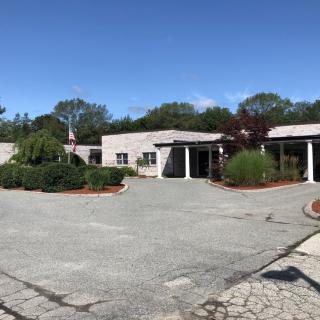 A single-story building with an adjacent parking lot and surrounding greenery under a clear blue sky.