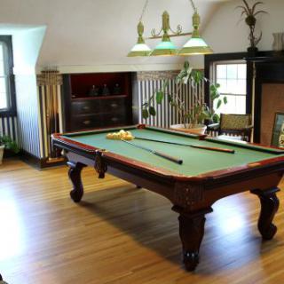 A cozy game room featuring a classic pool table with cues and balls, surrounded by wooden flooring and illuminated by an overhead light.