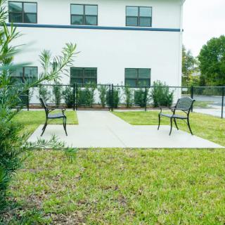 A small patio area with chairs, surrounded by grass and a white building in the background.