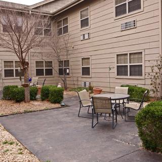 A cozy outdoor patio area with a table and chairs, surrounded by shrubs and adjacent to a building.