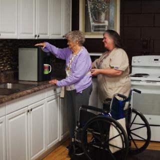 A caregiver assists an elderly woman in the kitchen while a wheelchair is nearby, highlighting the importance of support and independence.