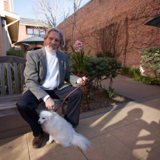 A man sitting on a bench in a garden with a small white dog.