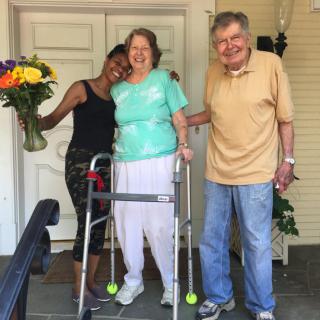 An elderly couple is greeted by a young woman holding a bouquet of flowers on their porch, all smiling happily.