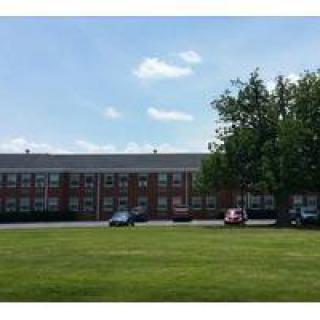 A school building with a grassy lawn, trees, and parked cars under a clear blue sky.