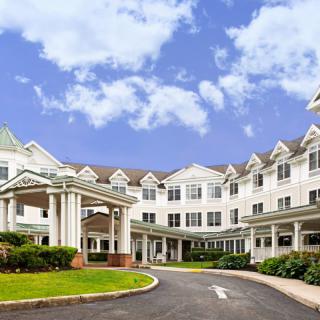 A large residential building with a covered entrance and surrounding greenery under a partly cloudy sky.