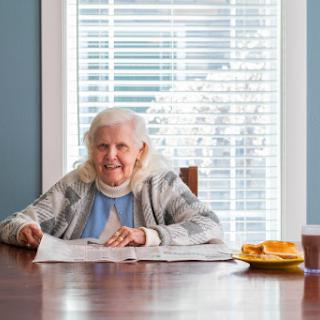An elderly woman enjoys reading a newspaper at the dining table with snacks beside her.