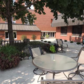 A serene outdoor patio area featuring a round glass table surrounded by chairs, complemented by lush greenery and a brick building backdrop.
