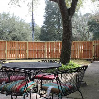 A cozy backyard patio with a metal table and chairs, accented by colorful cushions, under the shade of a large tree.