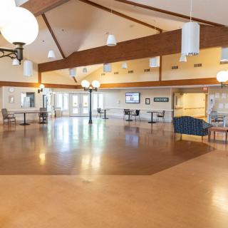 A spacious community hall with wooden flooring and bright lighting, featuring seating areas and windows along the walls.