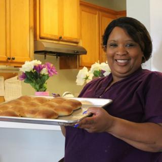 A woman smiling in a kitchen holding a tray of freshly baked bread with flowers in the background.