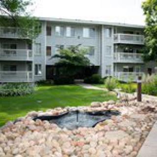 A three-story apartment building with landscaped gardens and a small pond in the foreground.
