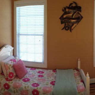 A cozy bedroom with floral bedding and pink pillows, illuminated by natural daylight through window blinds.