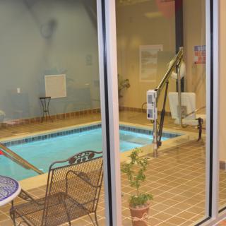 Indoor swimming pool area with chairs, tables, and a pool lift visible through a glass window.