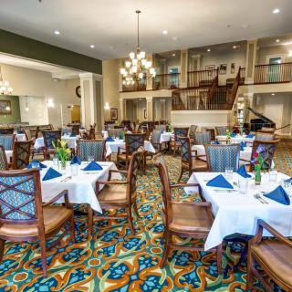 A spacious and elegantly decorated dining room with neatly arranged tables and chairs, featuring chandeliers and a vibrant carpet design.