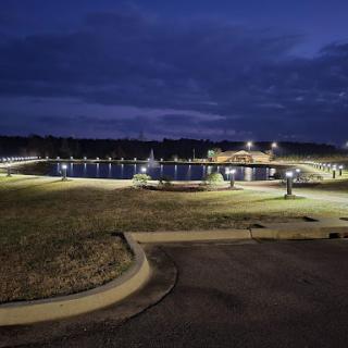 A serene view of a park by the water at night, illuminated by soft lights against a dusky sky.