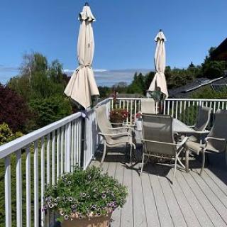 A sunny outdoor deck with a table, chairs, umbrellas, and potted plants overlooking a lush landscape.