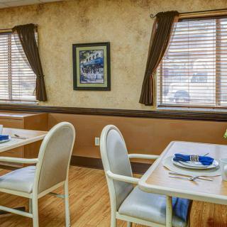 A cozy dining area with neatly arranged tables and chairs, decorated with a painting on the wall and windows with curtains and blinds.