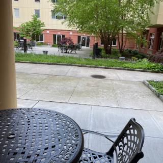 A quiet courtyard scene featuring a table and chairs surrounded by greenery and nearby buildings.