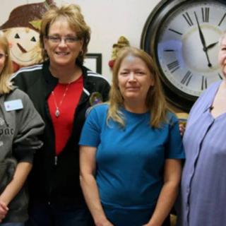 A group of four people standing indoors, with a large clock on the wall behind them.