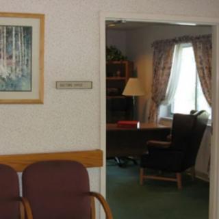 A view of an office through a waiting room, featuring chairs, a desk, and a window with curtains.