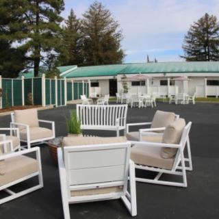 An outdoor seating area with white chairs and a table set on a patio, surrounded by trees and adjacent to a building.