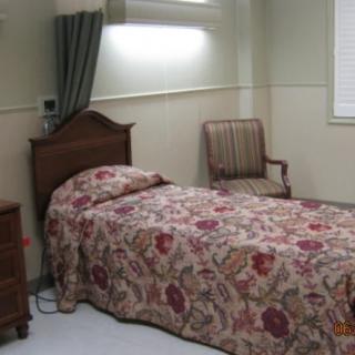 A simple bedroom with a single bed covered in a floral bedspread, accompanied by an armchair and nightstand.