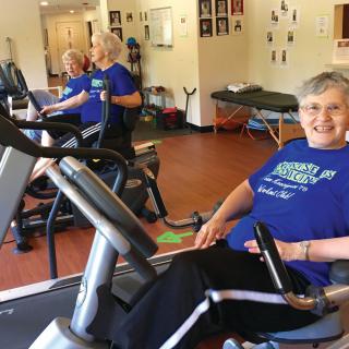An elderly woman smiles while exercising on a stationary bike in a fitness center.