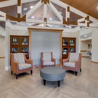 A modern lobby with upholstered chairs and a round table, framed by bookshelves and decorative beams.